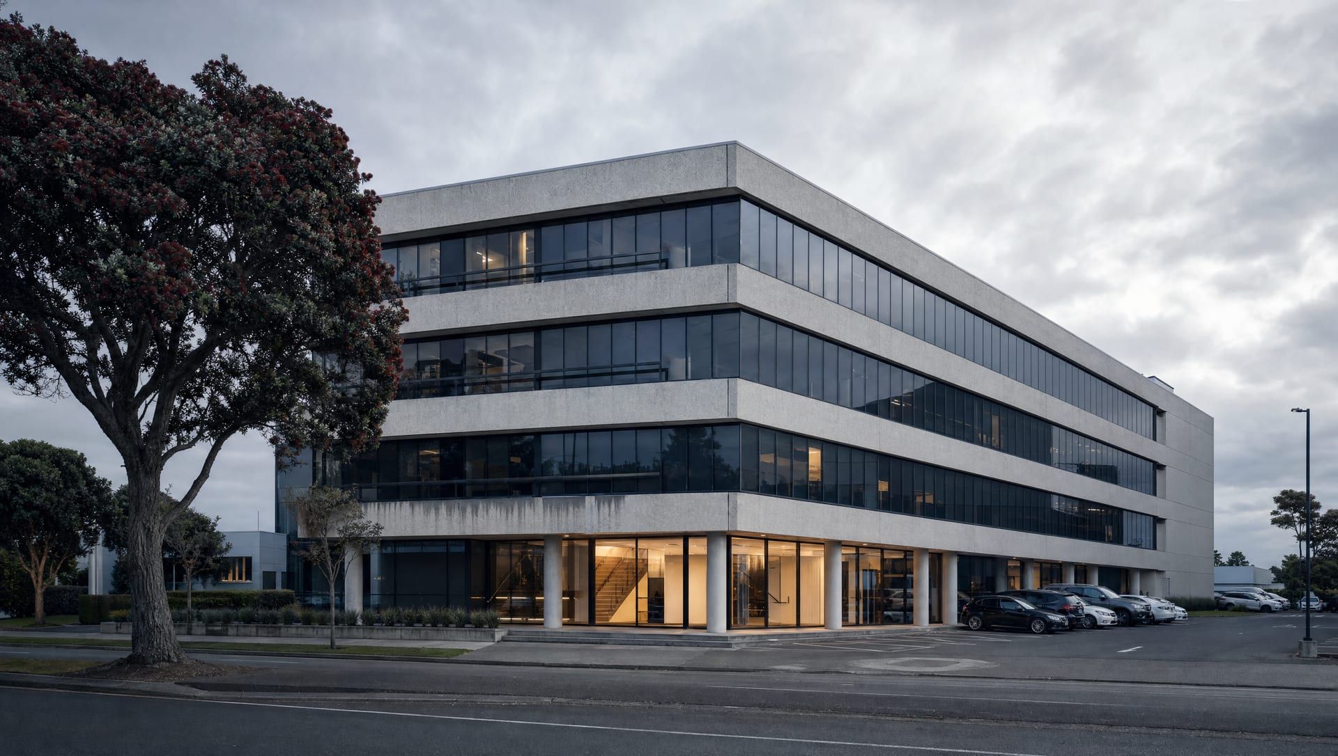 A four-storey precast concrete office building on a New Zealand business-park street under an overcast sky, its lobby lit.