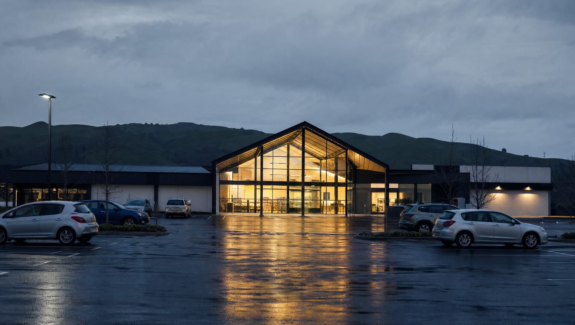 A single-storey suburban shopping centre at dusk, its tall glazed entrance lit, with a wet car park in the foreground.