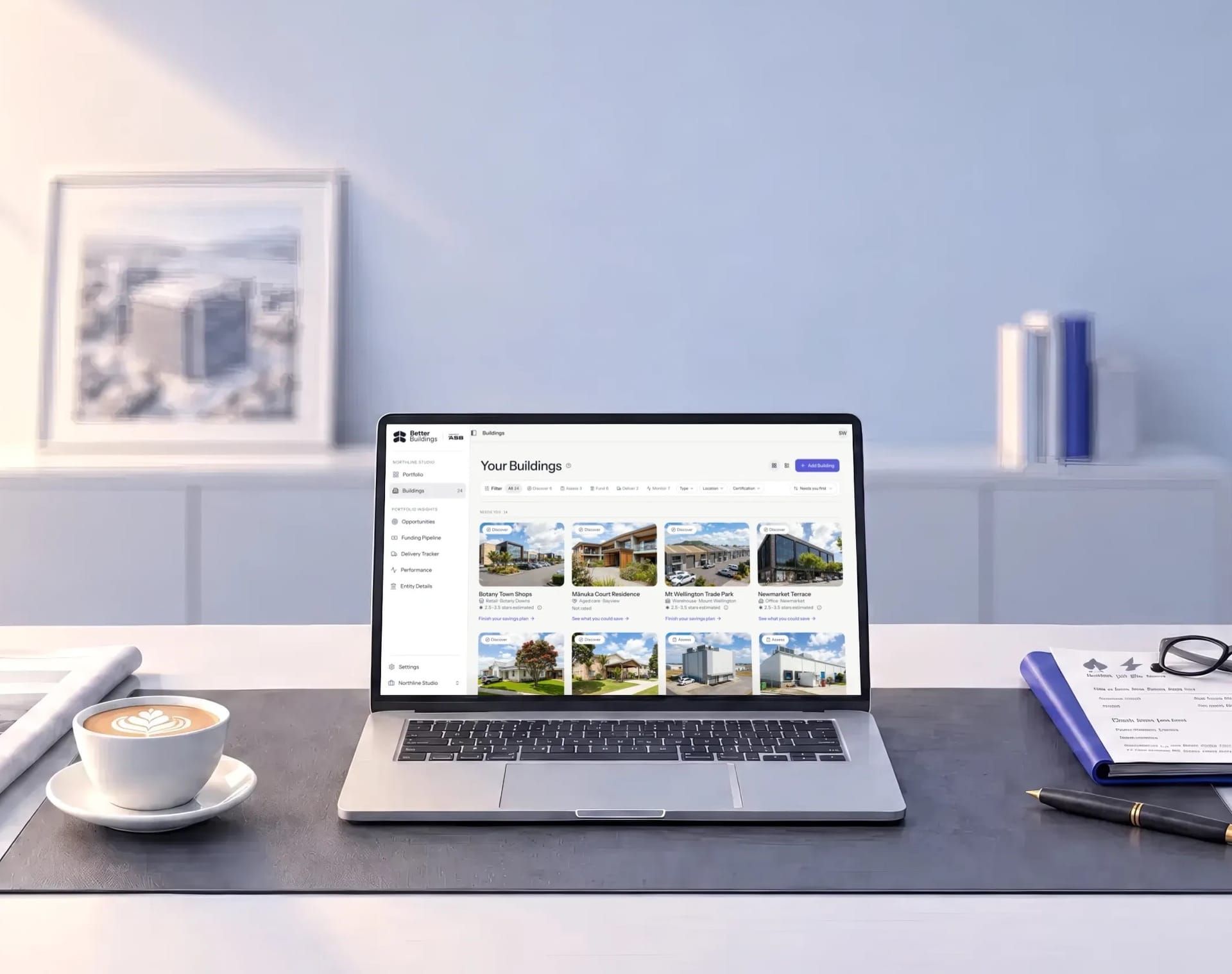 An office desk by a window over the city, with the Better Buildings app open on a laptop showing a portfolio of buildings.